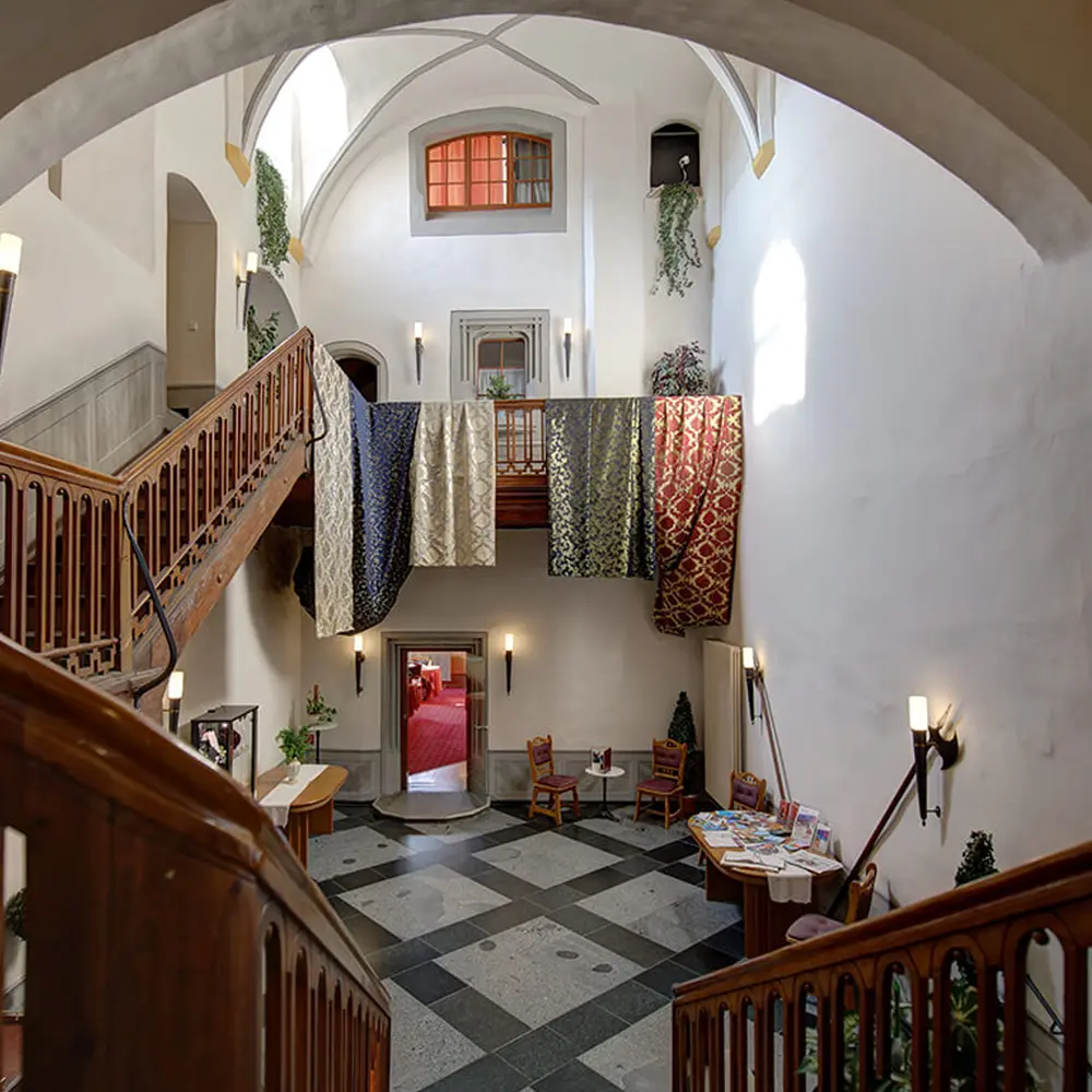 Blick von der Treppe hinab in die Eingangshalle mit schwarz-weißem Steinboden, Wandleuchten und den Tüchern an der Galerie.