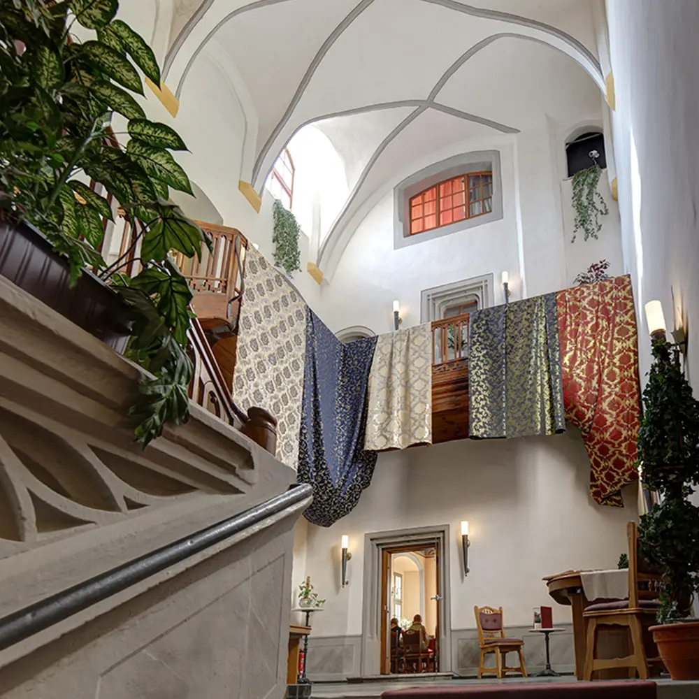 Die hohe Halle des Frenzelhofs: weiße Gewölbe, eine Holztreppe zur Galerie, über deren Brüstung Damaststoffe in Blau, Gold und Rot hängen.