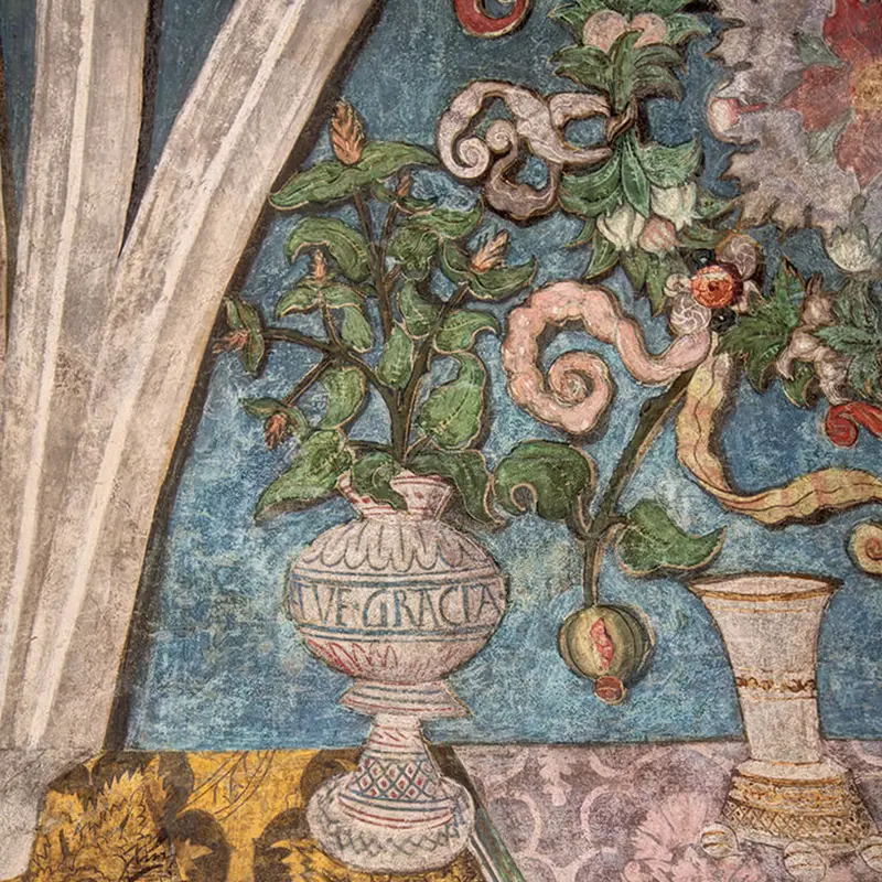 Detail der Malerei: eine Vase mit Inschrift, aus der Ranken mit Blüten wachsen, vor blauem Grund.