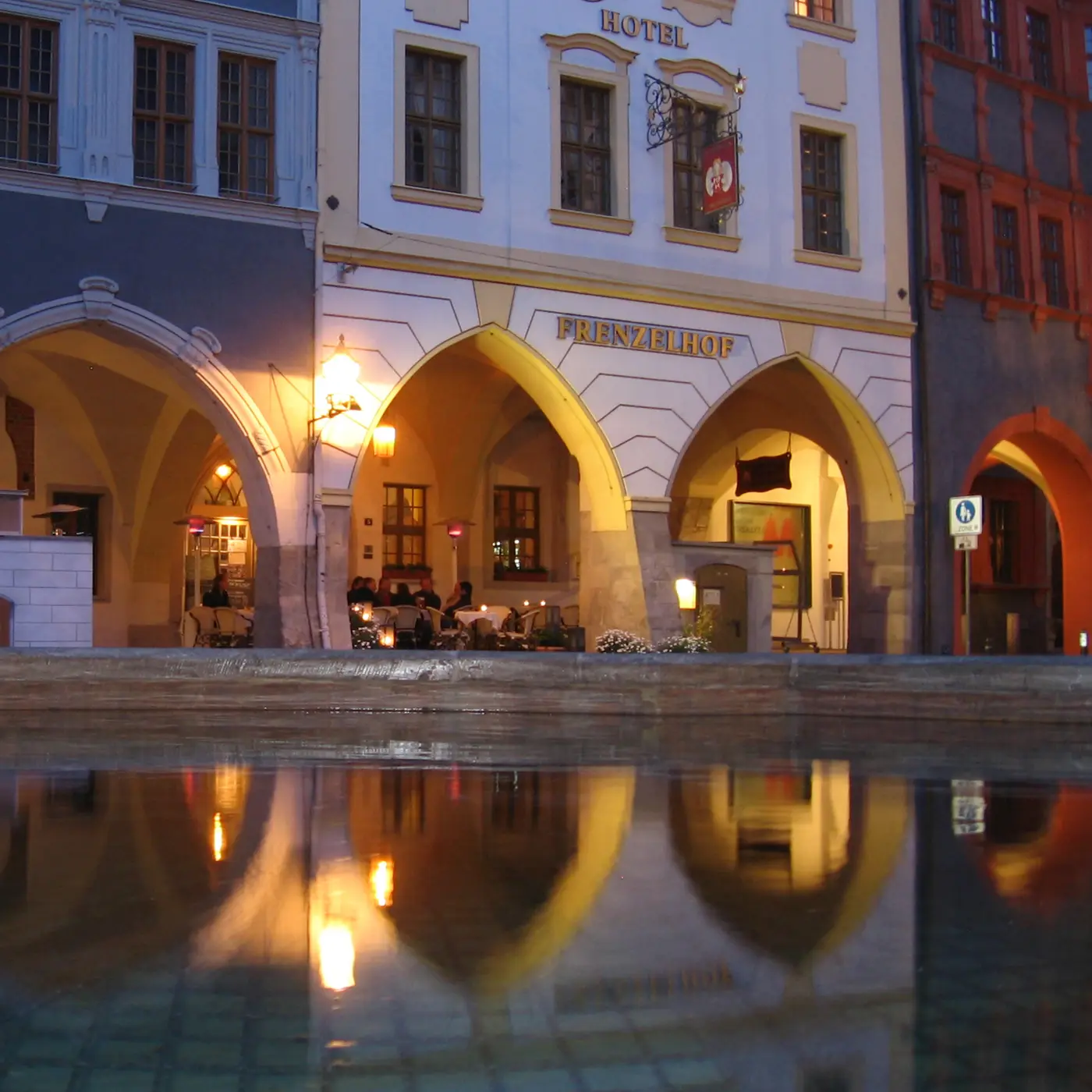 Die Fassade des Frenzelhofs am Abend: drei erleuchtete Spitzbogen-Lauben mit dem Schriftzug Frenzelhof, gespiegelt in einer Wasserfläche.