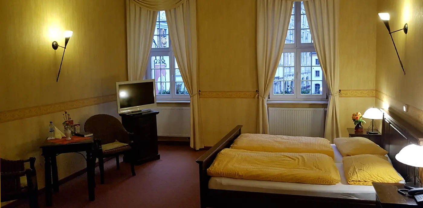 Ein Appartement mit warm gelben Wänden, Doppelbett, Sitzecke und zwei hohen Fenstern mit Vorhängen.