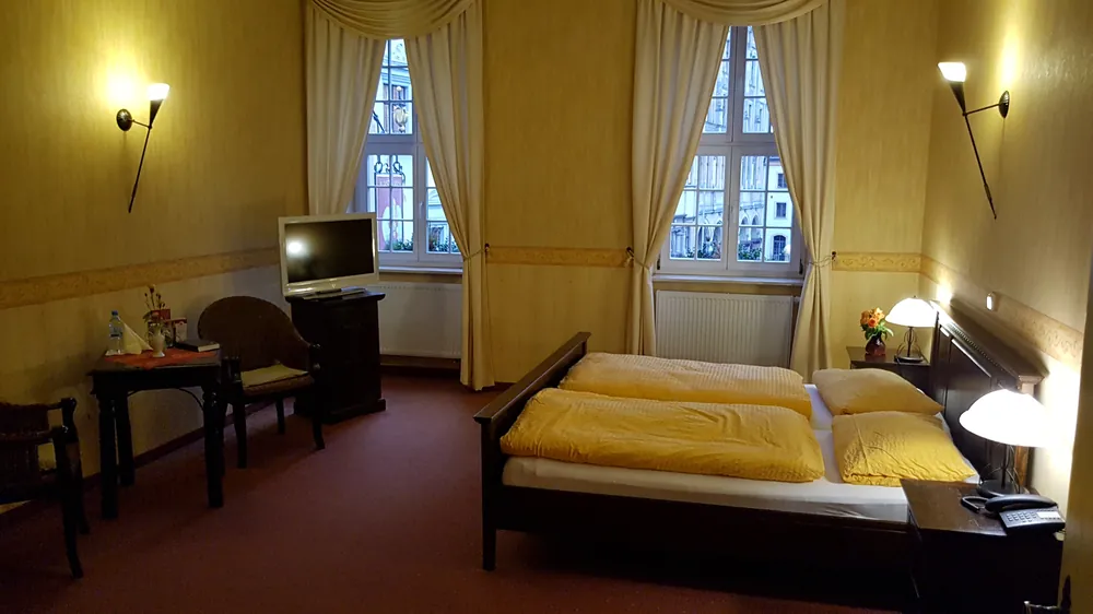 Ein Appartement mit gelb gestrichenen Wänden, Doppelbett mit gelber Wäsche, Wandleuchten, Fernseher und zwei hohen Fenstern.