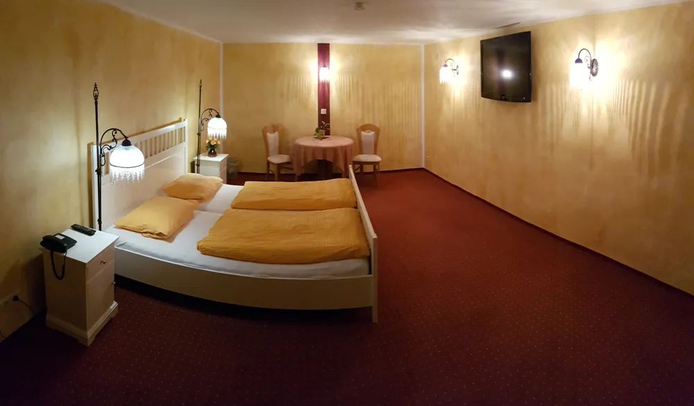 Großes Appartement mit weißem Doppelbett, Nachttisch mit Telefon, Sitzgruppe und Fernseher an der Wand.